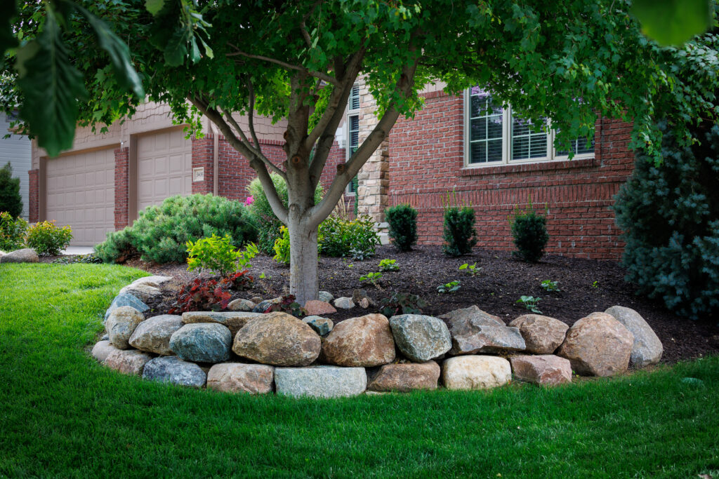 A well-landscaped front yard featuring a mature tree, stone border, and various plants and shrubs against a brick house.