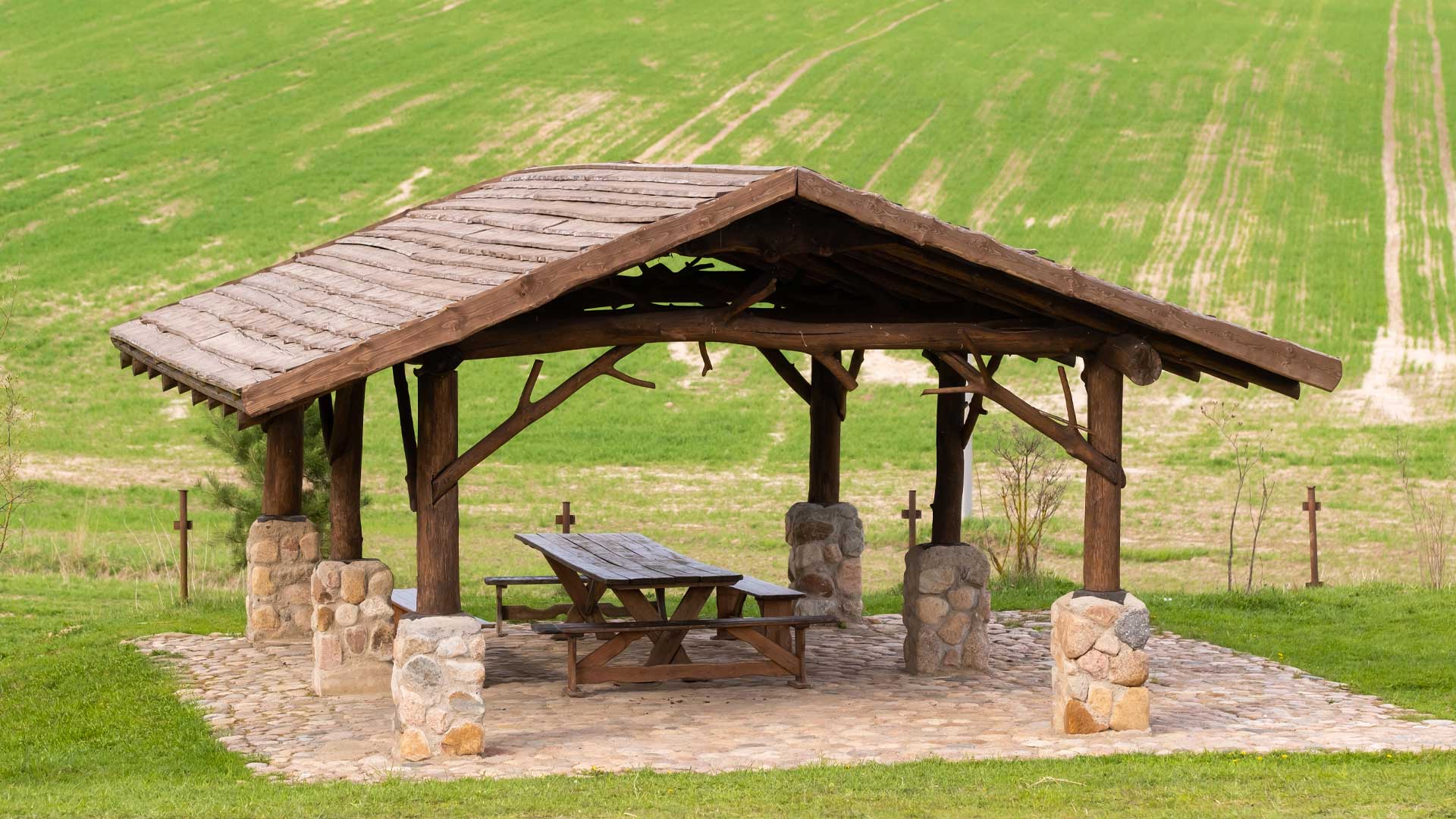 A wooden gazebo with a gabled roof stands on a cobblestone foundation in a green field, featuring a picnic table underneath.