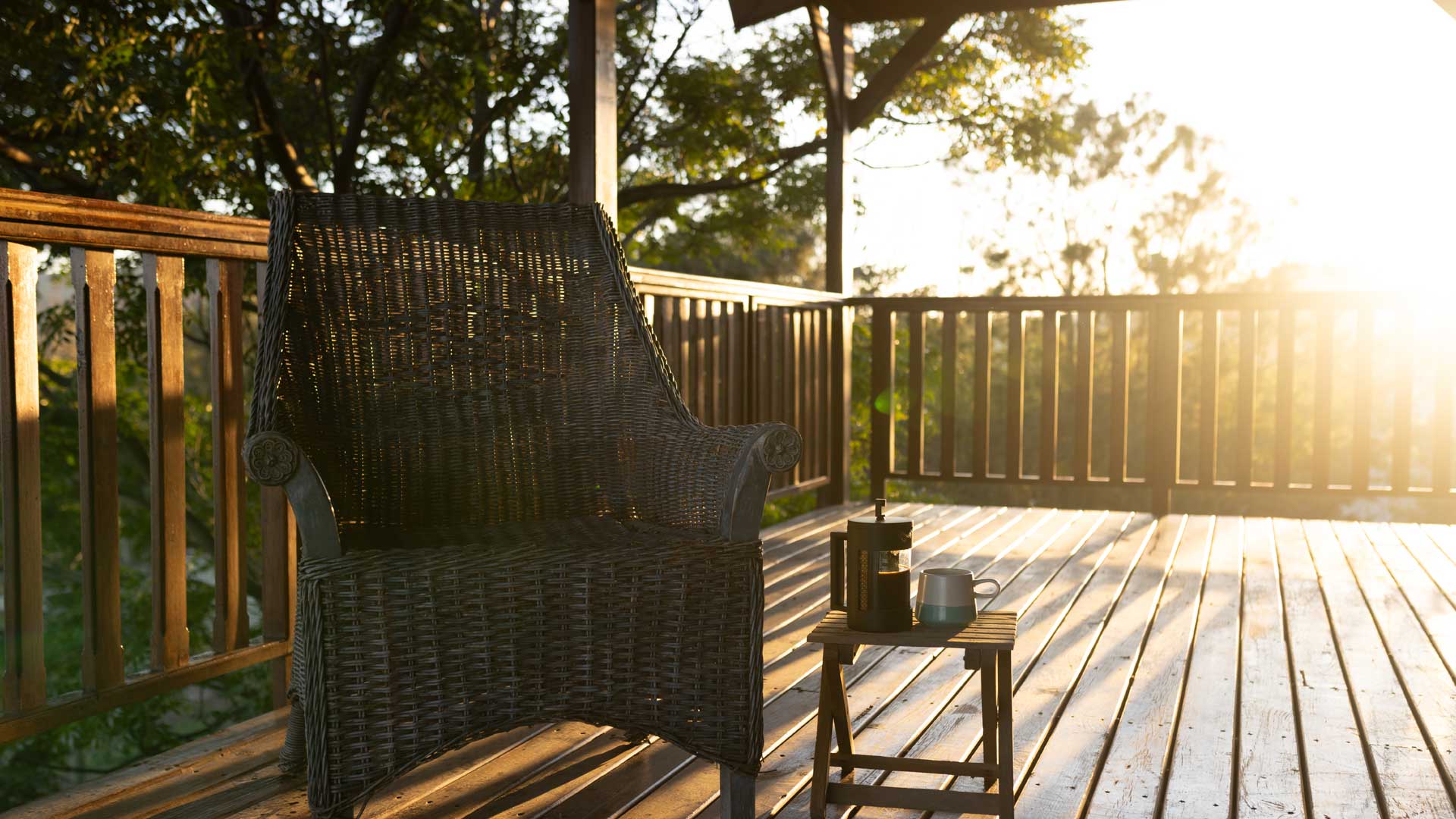 A cozy armchair on a cedar deck with a small table holding a French press and a mug, illuminated by the warm glow of the setting sun.