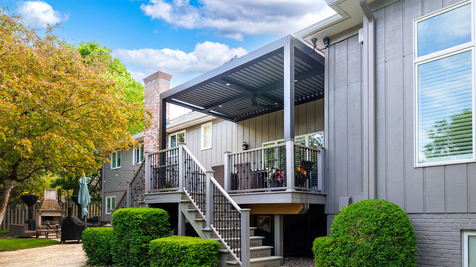 A modern deck with a SruXure pergola, featuring stylish railings, stairs, and a cozy outdoor seating area, attached to a two-story house with grey siding.