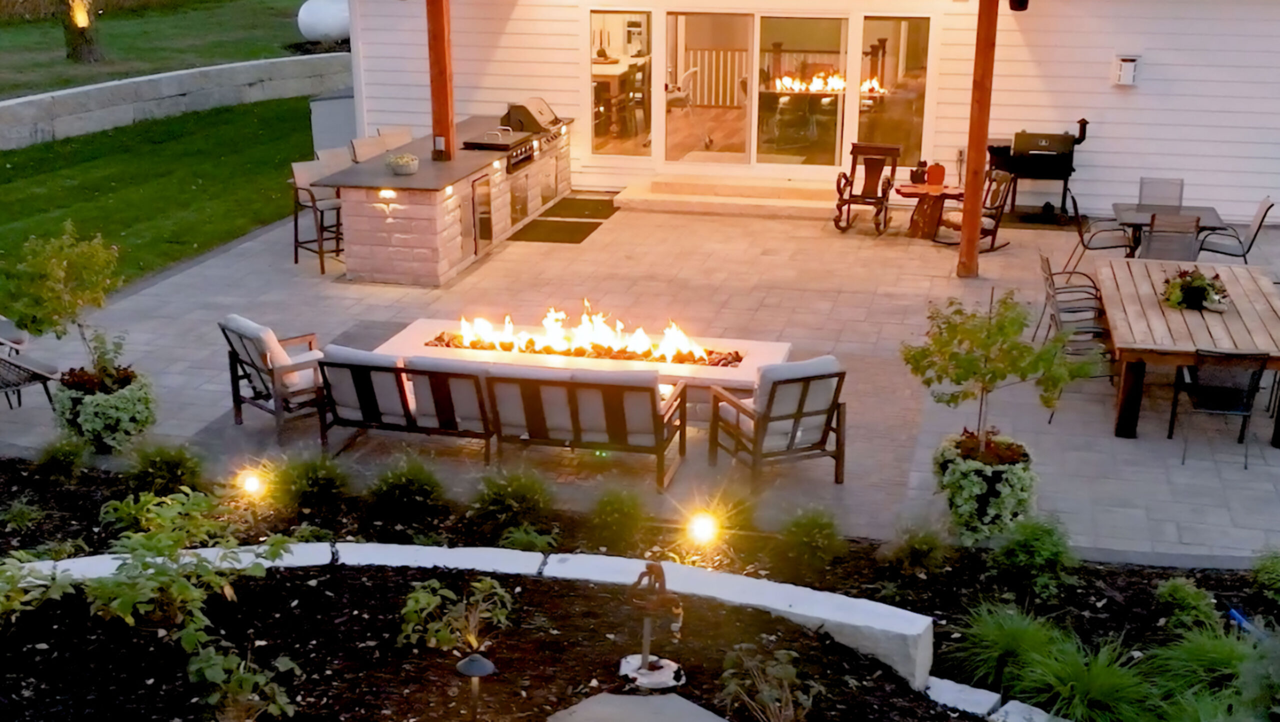 Evening view of a backyard patio with a fire feature, outdoor kitchen, and seating area