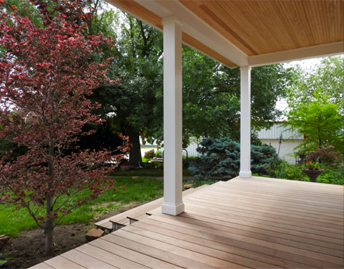 A wooden deck with white columns, overlooking a landscaped backyard with trees and plants.