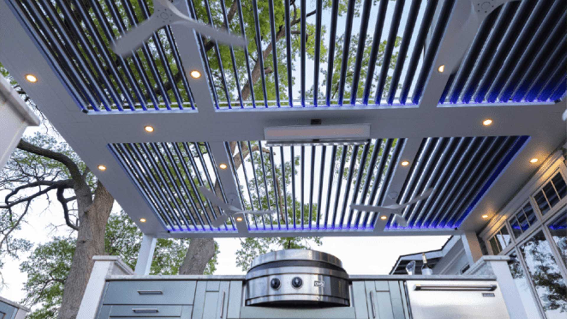 A modern SruXure pergola with adjustable louvers, integrated lighting, and ceiling fans, covering an outdoor kitchen area.