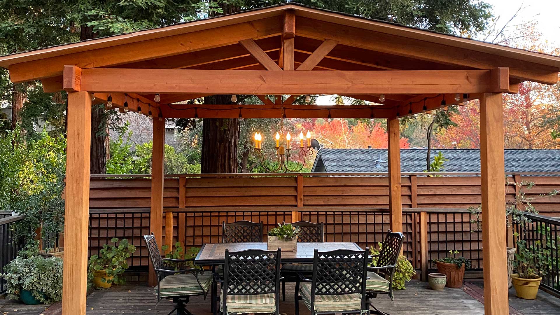 A well-crafted wooden pergola with a gabled roof stands on a backyard deck, surrounded by lush greenery. Underneath the pergola, a dining table with six chairs is set, ready for an outdoor gathering.