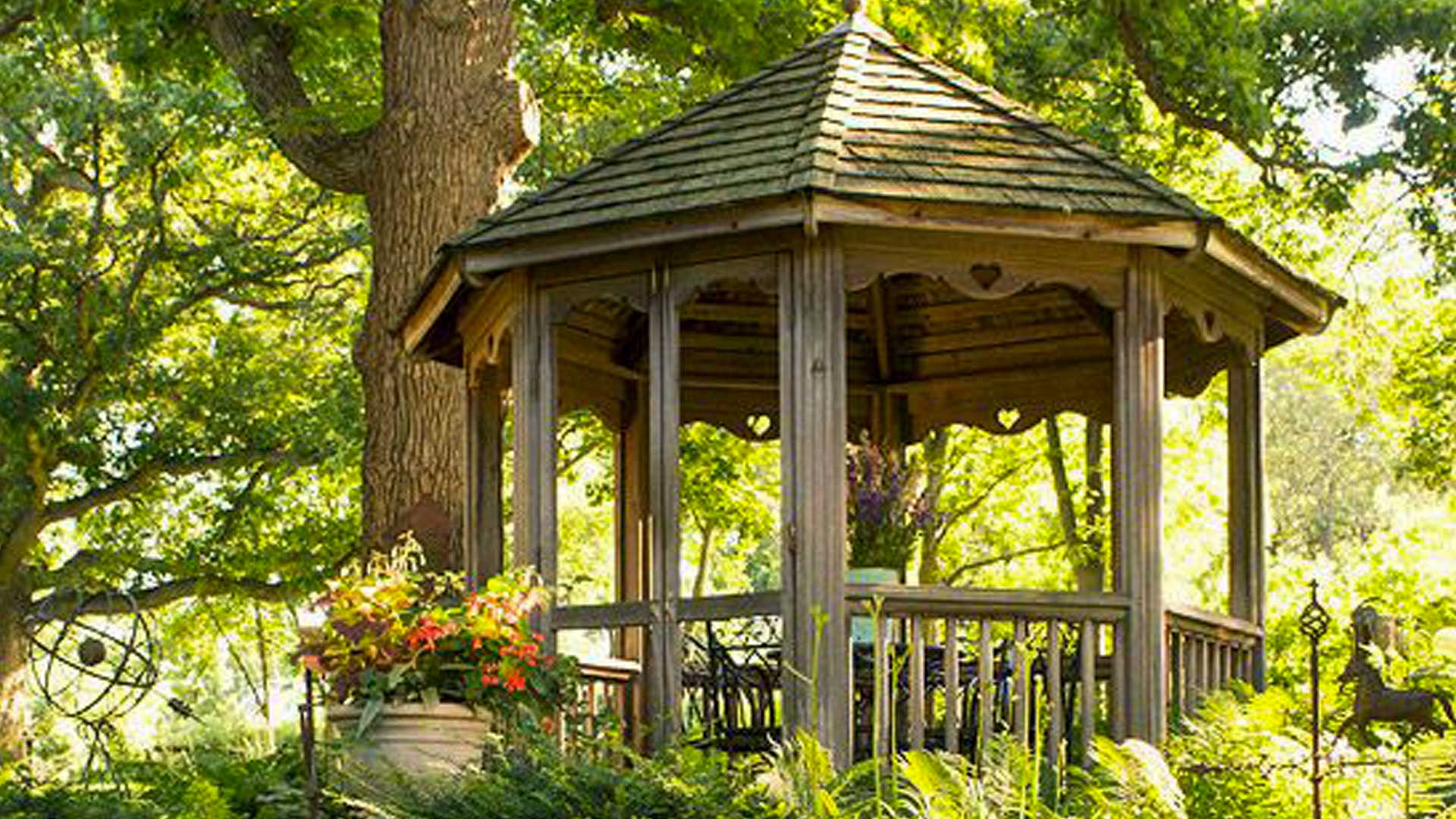 A charming wooden gazebo nestled in a lush garden with vibrant flowers and greenery, surrounded by mature trees.