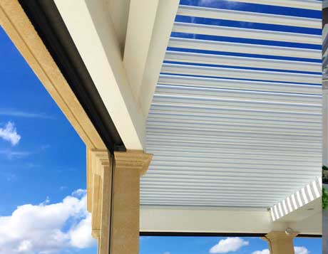 A SruXure pergola with adjustable white louvers, set against a bright blue sky with scattered clouds.