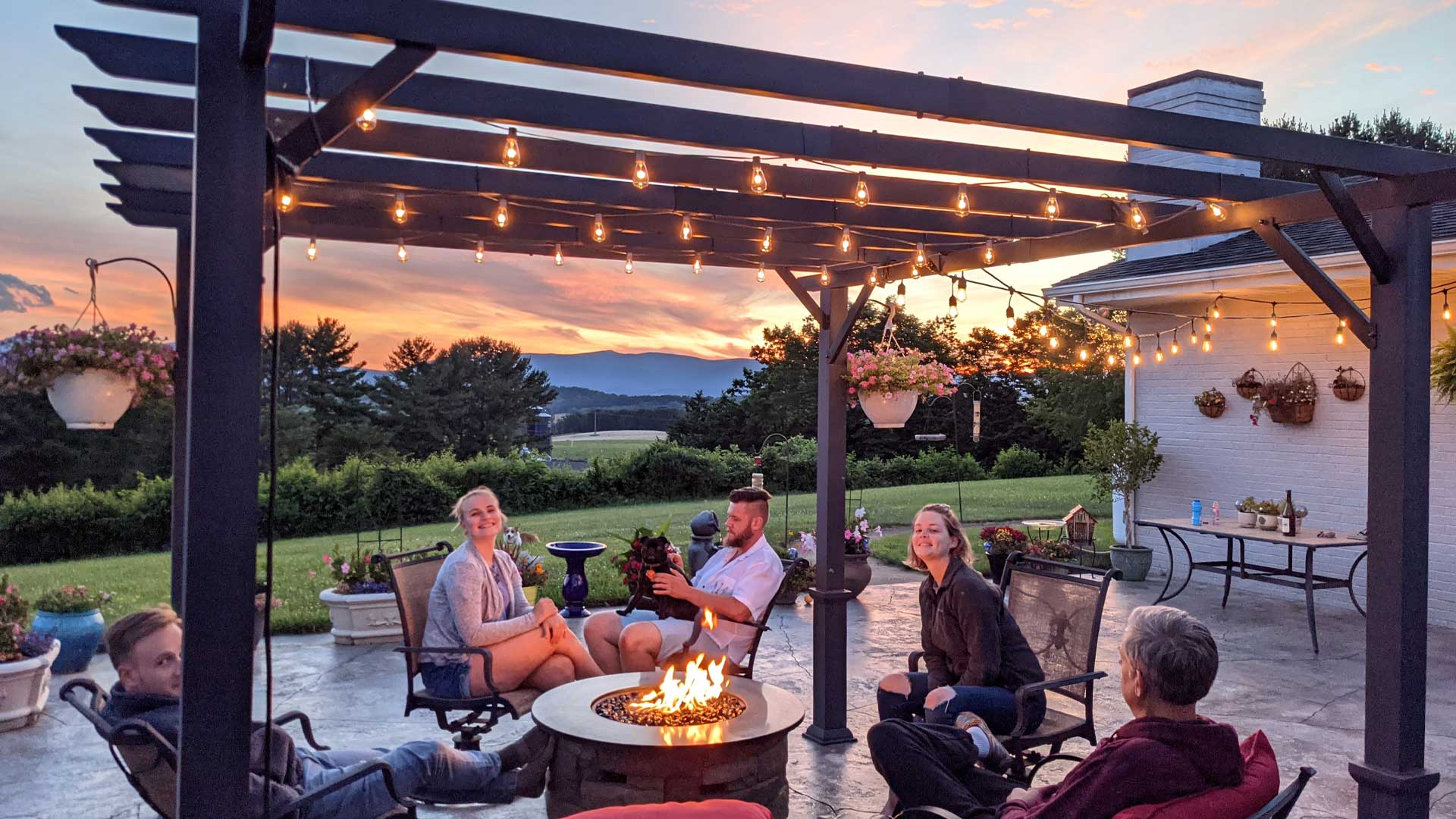 A family relaxing around a fire table under a metal pergola with string lights at sunset, set in a spacious backyard.