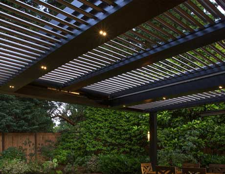 A modern SruXure pergola with adjustable louvers and integrated lighting, providing shade over an outdoor seating area in a lush backyard.