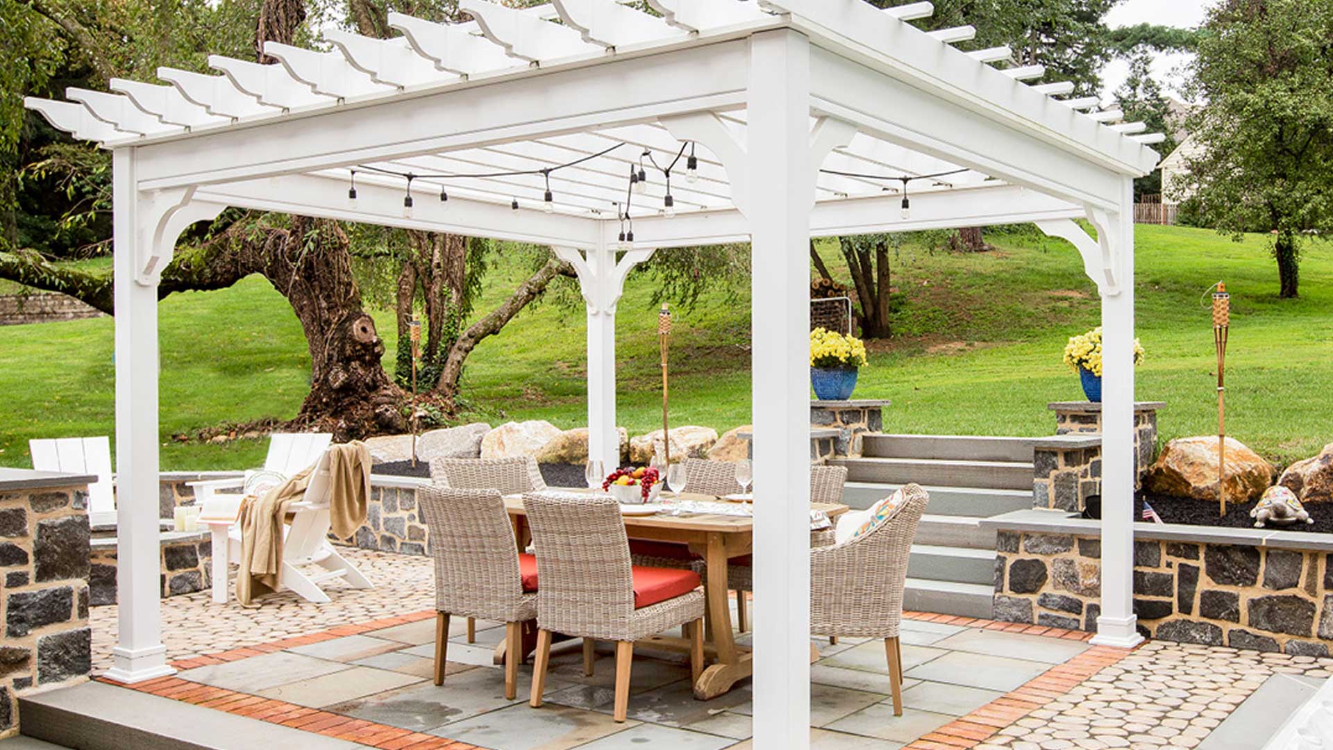 A stylish white vinyl pergola with string lights stands over an outdoor dining area, featuring a wooden table and wicker chairs, set in a landscaped backyard with stone patio and steps.