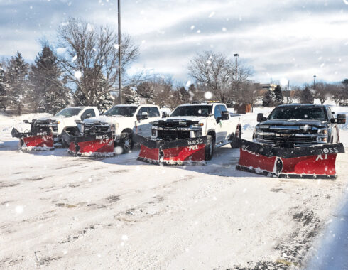 A fleet of snow plow trucks equipped with red plows, lined up in a snowy parking lot during a snowfall.